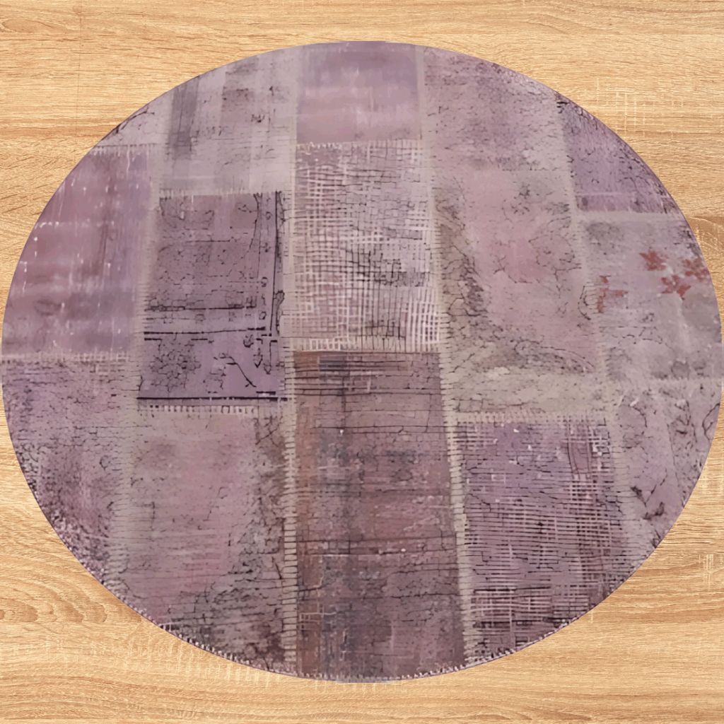 A round rug with a distressed, patchwork-style pattern in shades of mauve, lavender, and dusty rose. The rug is displayed on a light wood floor, showing its soft, muted tones.