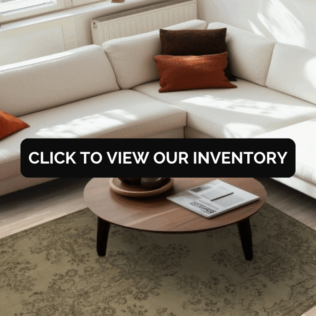 A bright living room with a white sectional sofa accented by rust-colored pillows. A round wooden coffee table rests on a soft olive-green patterned rug, with text overlay reading, “Click to view our inventory.”