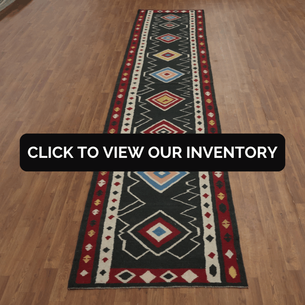 A long, narrow runner rug with a bold geometric pattern in black, red, white, yellow, and blue displayed on a wood floor. The center features a black overlay with white text reading, “Click to view our inventory.”