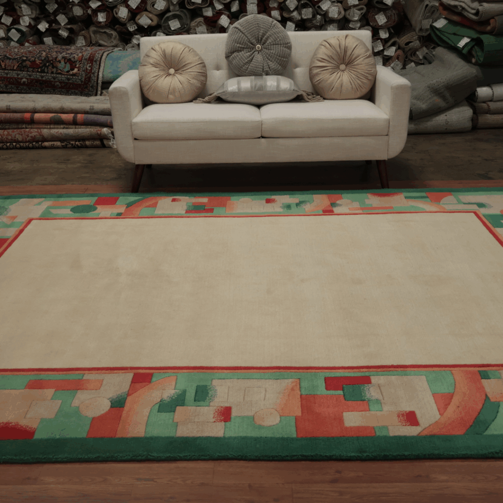 A beige sofa with round pillows sits in front of a display of rolled rugs, placed on a large cream rug bordered by colorful geometric designs in coral, mint, and peach.