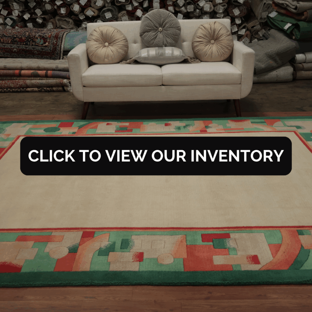 A cream sofa with round pillows sits atop a large beige rug bordered by abstract geometric patterns in green, red, and peach. Rolled rugs fill the background of the warehouse setting.
