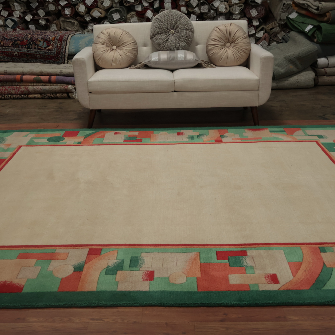 A beige sofa with round pillows sits in front of a display of rolled rugs, placed on a large cream rug bordered by colorful geometric designs in coral, mint, and peach.