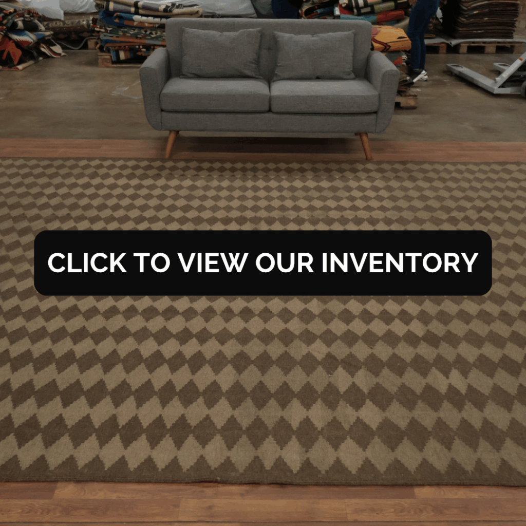 A gray sofa sits on a large brown rug with a diamond pattern in a warehouse filled with stacked rolled rugs. The text overlay reads, “Click to view our inventory.”
