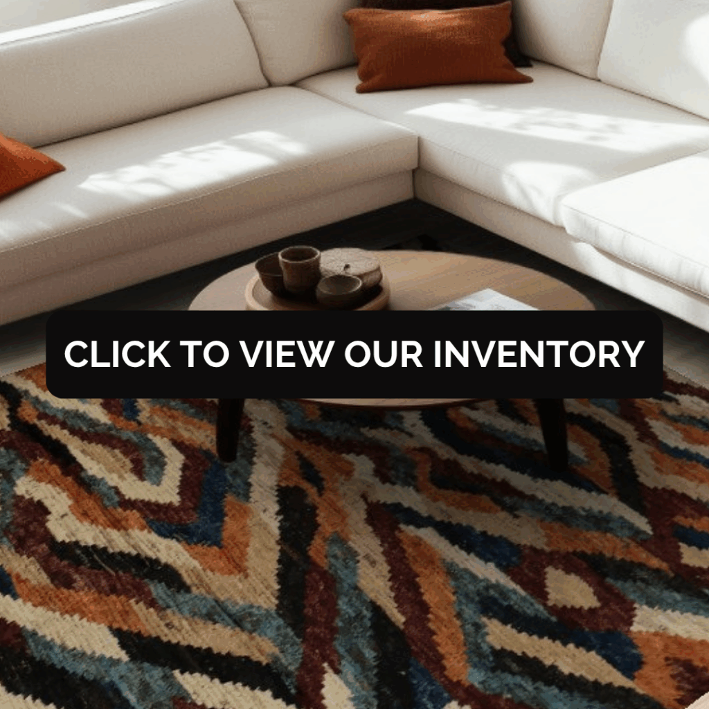 A cozy modern living room with a cream sectional sofa and burnt orange throw pillows. A round wooden coffee table holds small bowls and a magazine, centered over a bold geometric rug in shades of rust, navy, gold, and cream. Text overlay reads “Click to View Our Inventory.”
