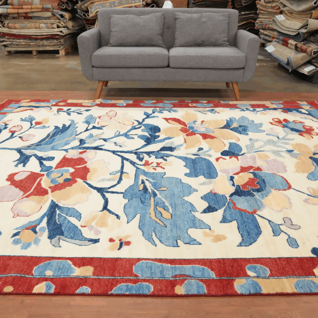 A gray mid-century sofa sits on a large cream rug with a bold floral pattern in shades of blue, red, and gold. Rolled rugs line the background of a showroom. Text overlay reads “Click to View Our Inventory.”