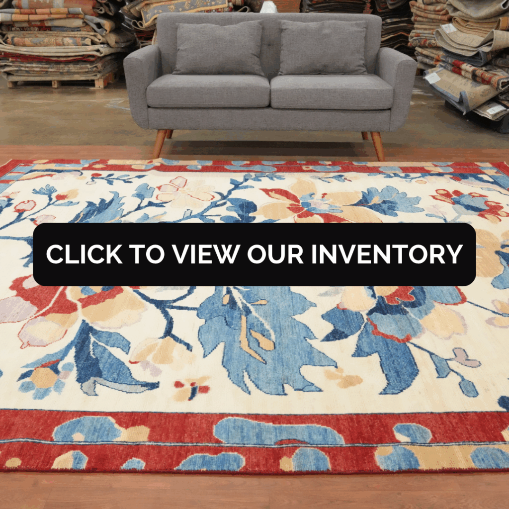 A vibrant floral rug with red and blue leaves and flowers covers the floor in front of a gray sofa. The background shows stacks of rolled rugs in a showroom setting.
