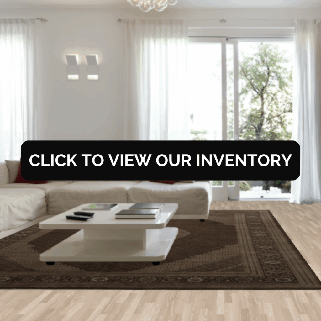 A bright, minimalist living room with a cream sectional sofa, red accent pillows, and a sleek white coffee table. A dark brown patterned rug anchors the space, complemented by light wood floors and large windows letting in natural light. Text overlay reads “Click to View Our Inventory.”
